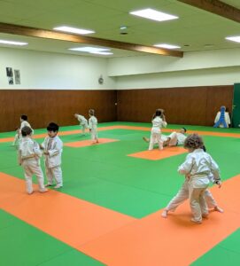 Une dizaine d’enfants, vêtus de kimonos blancs (judogis), pratiquent le judo dans un dojo spacieux et lumineux. Certains sont en train de s’échauffer ou de discuter en petits groupes, tandis que d’autres s’entraînent sur des tatamis verts et orange.