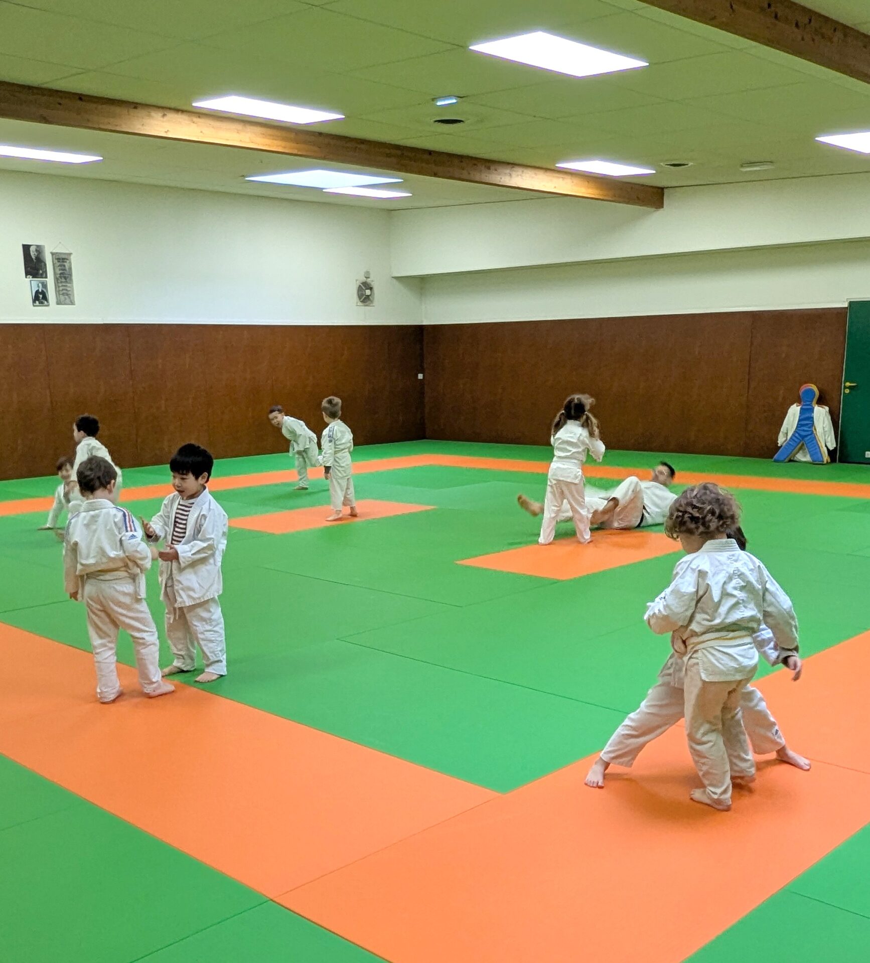 Une dizaine d’enfants, vêtus de kimonos blancs (judogis), pratiquent le judo dans un dojo spacieux et lumineux. Certains sont en train de s’échauffer ou de discuter en petits groupes, tandis que d’autres s’entraînent sur des tatamis verts et orange.